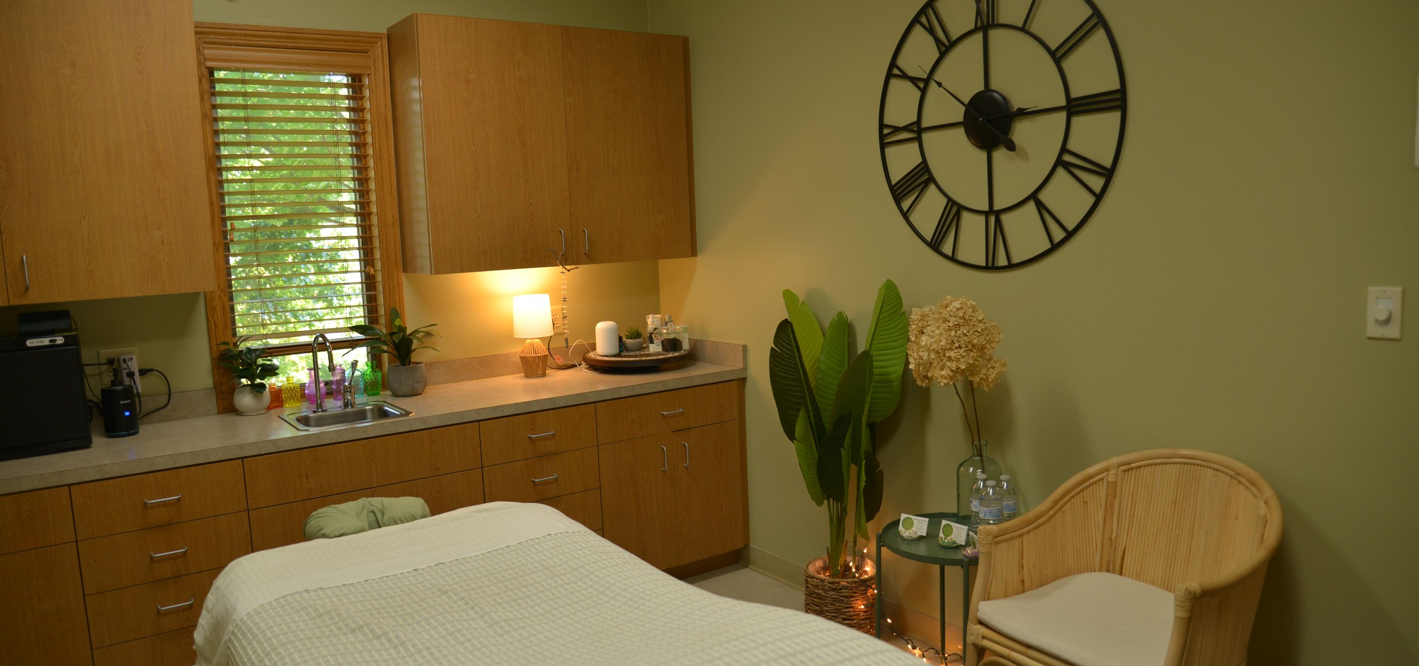 The sage-green treatment room with a massage table, plants and a large wall clock